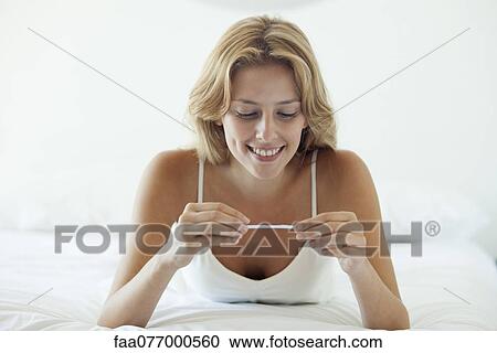 Stock Image - Young woman lying on bed looking at pregnancy test, smiling. Fotosearch