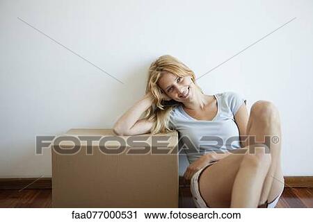 Stock Image - Young woman sitting on floor, leaning on cardboard box. Fotosearch