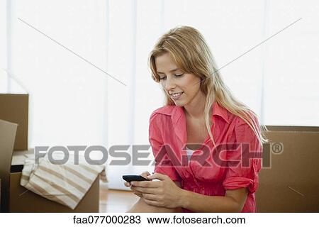 Stock Image - Young woman text messaging in new house, surrounded by cardboard boxes. Fotosearch