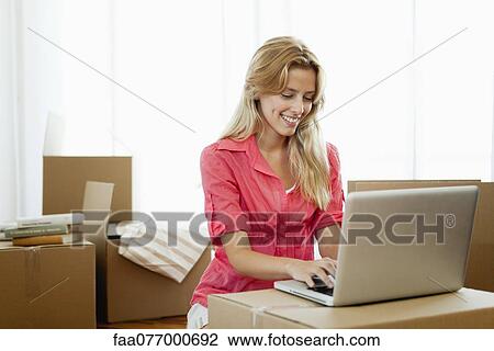 Stock Image - Young woman using laptop computer while surrounded by cardboard boxes. Fotosearch