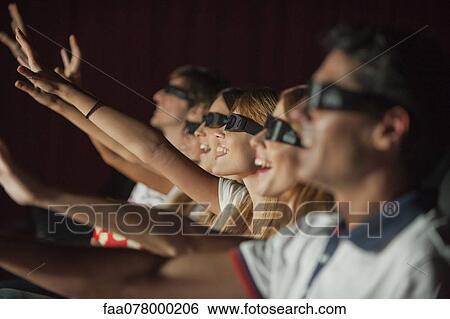 Stock Photograph - Audience wearing 3-D glasses in movie theater, arms reaching out. Fotosearch