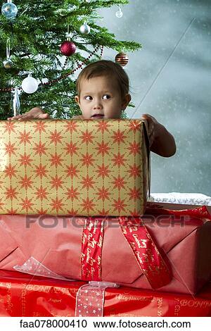Stock Image - Baby girl standing behind stack of Christmas gifts with mischievous expression. Fotosearch