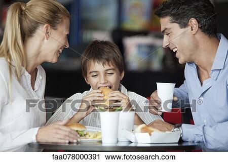 Stock Image - Boy eating hamburger in fast food restaurant with his parents. Fotosearch