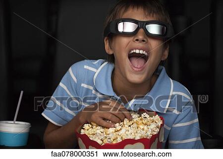 Stock Image - Boy laughing and eating popcorn during 3-D movie in theater. Fotosearch