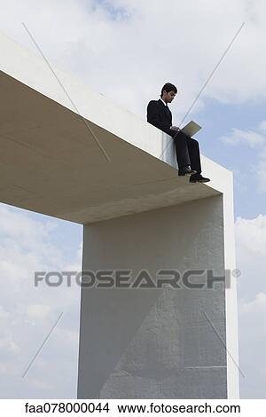 Picture - Businessman sitting on top of tall structure using laptop computer. Fotosearch