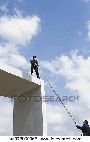 Businessman standing on top of tall structure, holding rope to help colleague View Large Photo Image Stock Photograph - Businessman standing on top of tall structure, holding rope to help colleague. Fotosearch
