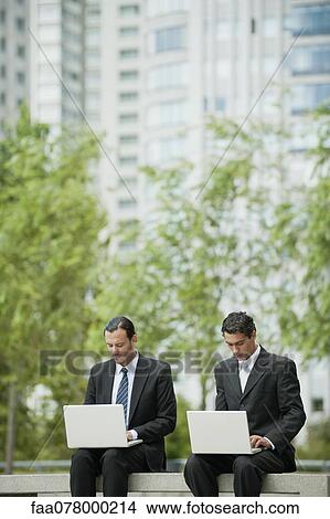 Picture - Businessmen sitting side by side using laptop computers outdoors. Fotosearch