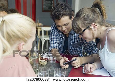 Picture - Friends hanging out together in cafe, looking at cell phone. Fotosearch