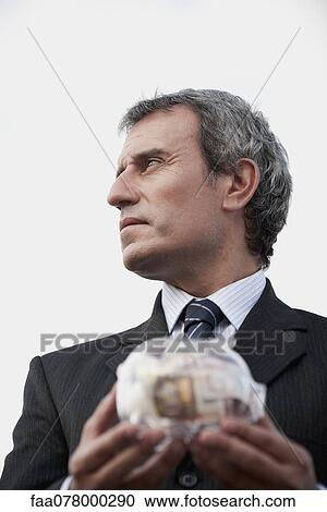 Stock Image - Mature man holding transparent piggy bank filled with euros. Fotosearch