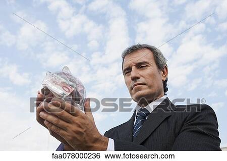 Stock Photograph - Mature man holding transparent piggy bank filled with euros. Fotosearch