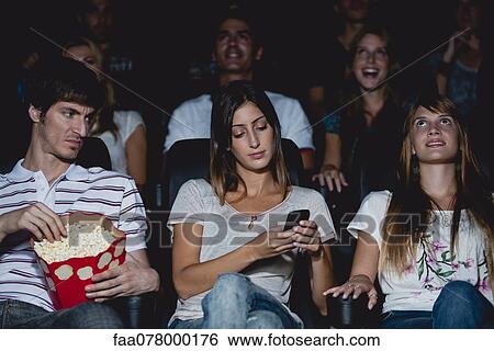 Stock Photograph - Woman using cell phone in movie theater, man looking over with annoyed expression. Fotosearch