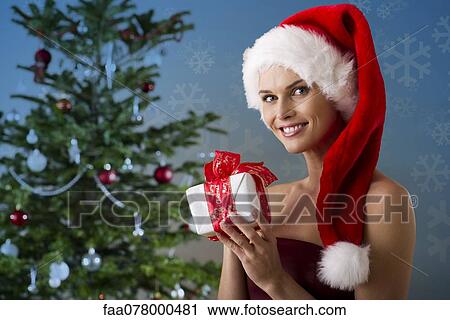 Stock Image - Woman wearing Santa hat and holding Christmas present, portrait. Fotosearch