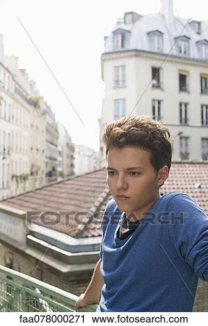Stock Image - Young man leaning against railing, looking away in thought. Fotosearch