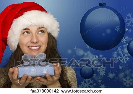 Stock Image - Young woman in Santa hat holding Christmas gift, portrait. Fotosearch