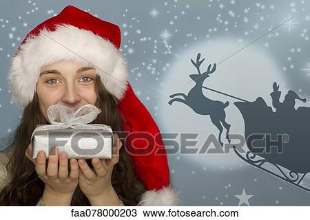Stock Image - Young woman in Santa hat holding Christmas present, graphic of Santa's sleigh in background. Fotosearch