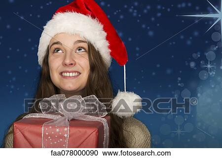 Stock Image - Young woman wearing Santa hat, holding Christmas present, portrait. Fotosearch