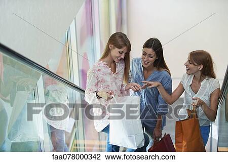 Stock Image - Young women discussing purchases while standing on escalator. Fotosearch