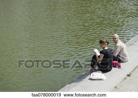 Stock Photo - Young women sitting beside lake together. Fotosearch