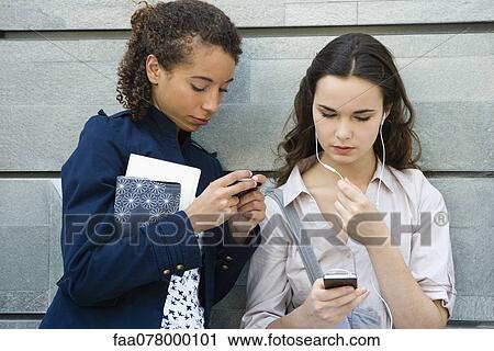 Stock Image - Young women together outdoors, text messaging and listening to MP3 player. Fotosearch