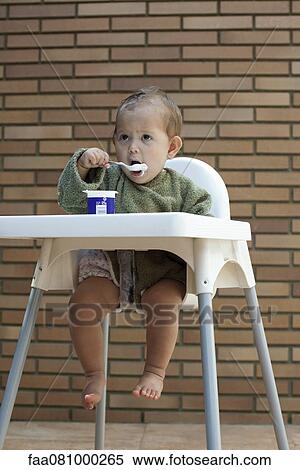 Stock Photography - Baby girl sitting in high chair, eating yogurt with spoon. Fotosearch