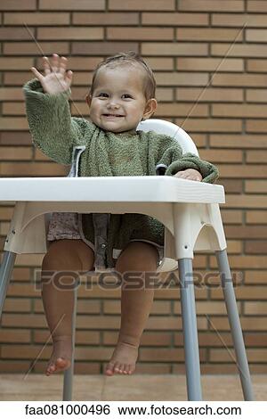 Stock Photograph - Baby girl sitting in high chair, waving. Fotosearch