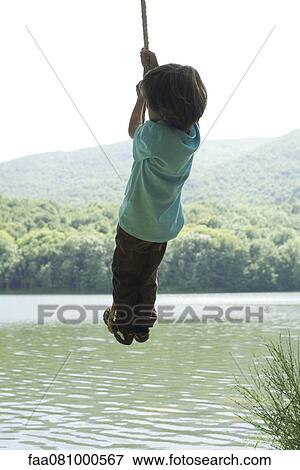 Stock Photo - Boy hanging from rope over river, rear view. Fotosearch