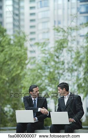 Business executives sitting outdoors with laptop computers, shaking hands View Large Photo Image Stock Photo - Business executives sitting outdoors with laptop computers, shaking hands. Fotosearch