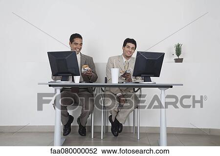 Stock Image - Businessmen eating fast food at desk in office. Fotosearch