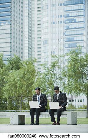 Businessmen sitting side by side using laptop computers outdoors View Large Photo Image Stock Photo - Businessmen sitting side by side using laptop computers outdoors. Fotosearch