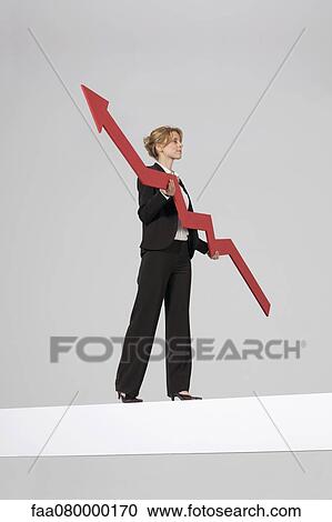 Stock Image - Businesswoman walking on balance beam, carrying large arrow indicating profits. Fotosearch