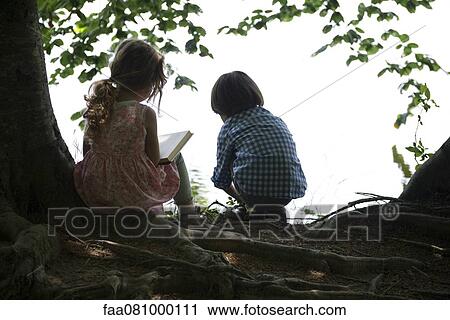 Stock Image - Children hanging out by lake, rear view, backlit. Fotosearch