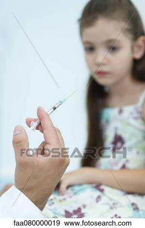 Stock Photo - Doctor's hand holding syringe, girl sitting in background. Fotosearch