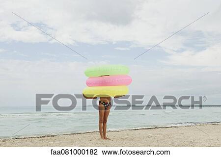 Stock Image - Girl holding stack of inflatable rings on beach, face obscured. Fotosearch