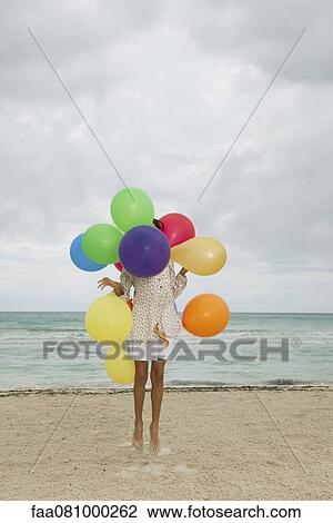 Stock Image - Girl jumping on beach, face obscured by bunch of colorful balloons. Fotosearch