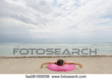 Stock Photograph - Girl lying on inflatable ring on beach, rear view. Fotosearch