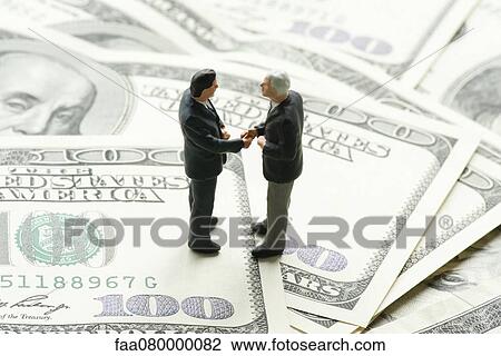 Stock Image - Male figurines shaking hands on pile of one hundred dollar bills. Fotosearch