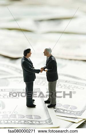 Stock Image - Male figurines shaking hands on pile of one hundred dollar bills. Fotosearch