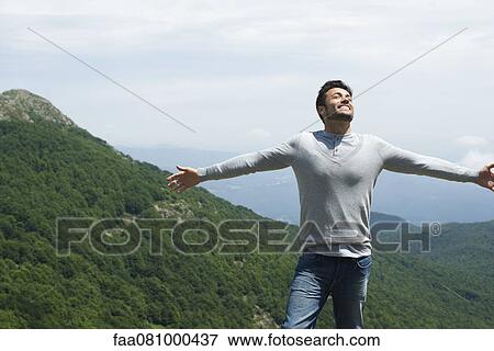Stock Photo - Mid-adult man with eyes closed and arms outstretched, mountainscape in background. Fotosearch