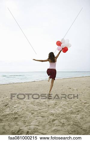 Stock Photography - Woman running on beach with bunch of balloons, rear view. Fotosearch