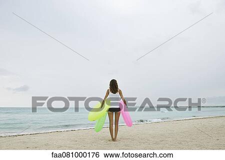Stock Photograph - Woman standing on beach, holding inflatable rings, rear view. Fotosearch