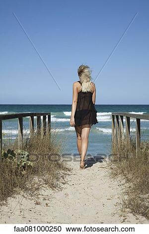 Stock Image - Woman walking on path toward sea, rear view. Fotosearch