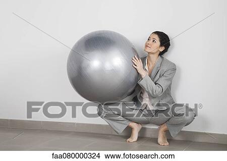 Picture - Young businesswoman crouching against wall holding fitness ball. Fotosearch