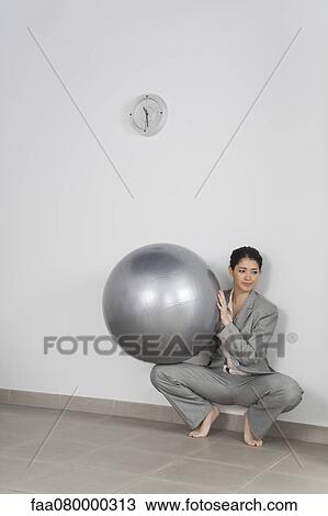 Stock Image - Young businesswoman crouching against wall holding fitness ball. Fotosearch