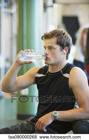 Young man drinking bottled water in gym room View Large Photo Image Stock Photo - Young man drinking bottled water in gym room. Fotosearch