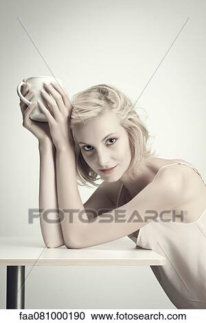 Stock Image - Young woman holding coffee cup at table, portrait. Fotosearch
