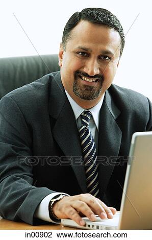 Stock Image - Businessman at desk, using laptop, looking at camera. Fotosearch