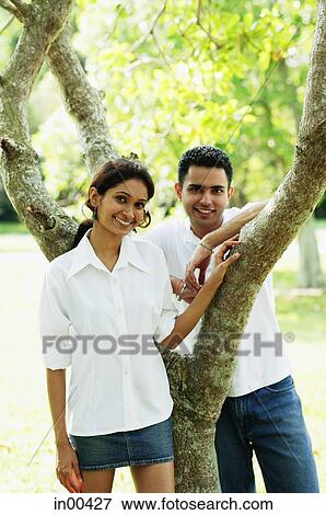 Stock Photo - Couple looking at camera, man leaning on tree. Fotosearch
