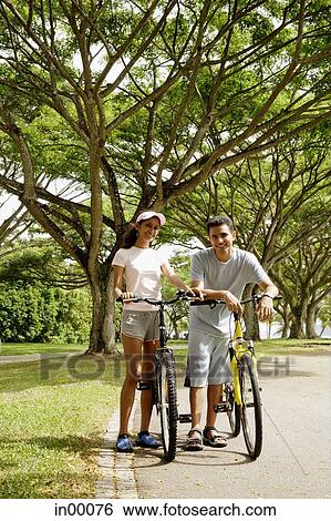 Couple standing with bicycles in park, looking at camera View Large Photo Image Stock Photograph - Couple standing with bicycles in park, looking at camera. Fotosearch