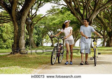 Stock Image - Couple walking in park, holding bicycles. Fotosearch