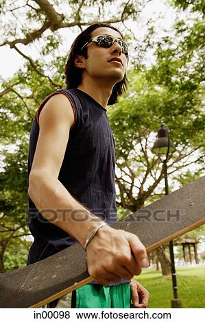 Stock Photo - Man wearing sunglasses, holding skateboard, low angle view. Fotosearch